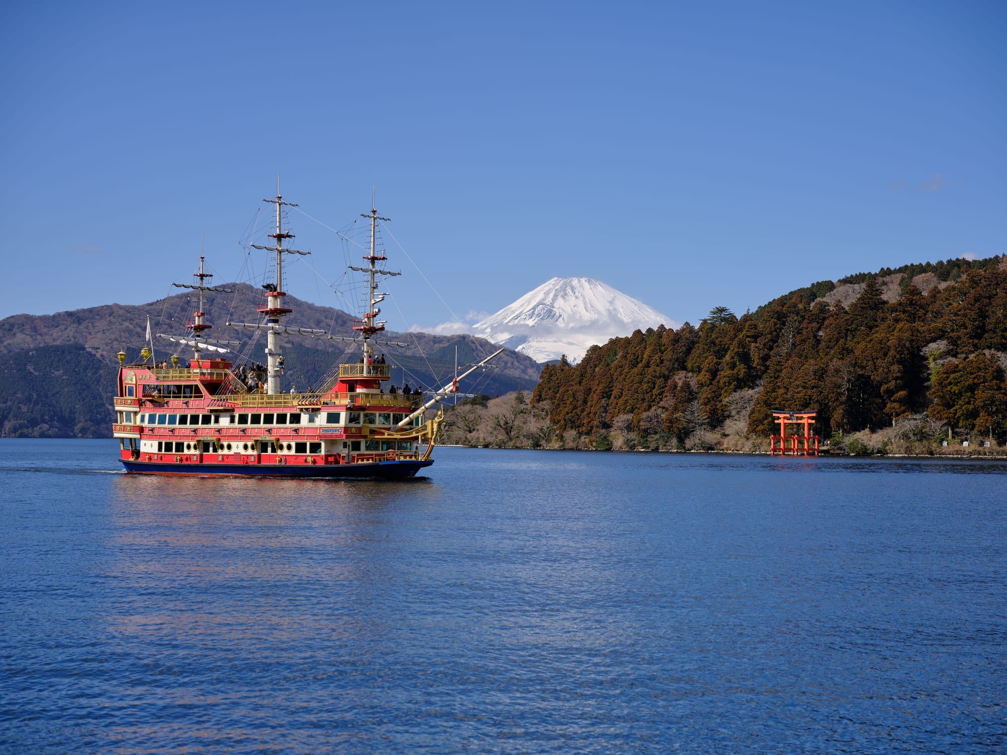 A colorful pirate ship sailing on Lake Ashi, with snow-capped Mount Fuji and a red torii gate on the shore