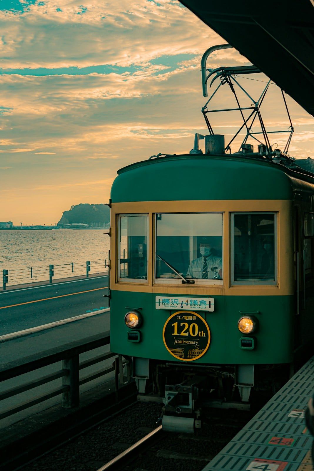 Green-and-cream Enoden train at a seaside platform at sunset, with the sea and Enoshima in the distance